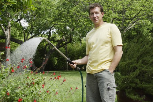 A man in a light yellow T-shirt and grey cargo shorts is standing in a lush, well-maintained garden in Fulham, holding a garden hose and watering red-flowered bushes in the foreground. Behind him, there is a neatly trimmed lawn with vibrant green grass, bordered by a mix of dense, neatly shaped evergreen shrubs and leafy trees with abundant foliage, providing shade and natural privacy. The garden surface appears to be a combination of rich soil bed and paved walkway, with a modern, slightly textured appearance. The background features tall trees with fresh green leaves, indicating the season is late spring or early summer, and the overall scene is well-lit with natural daylight, reflecting typical outdoor maintenance activities. This setting highlights the use of garden watering tools and landscaping efforts by Gardening Services Fulham to maintain a healthy and visually appealing outdoor space in the Fulham area.
